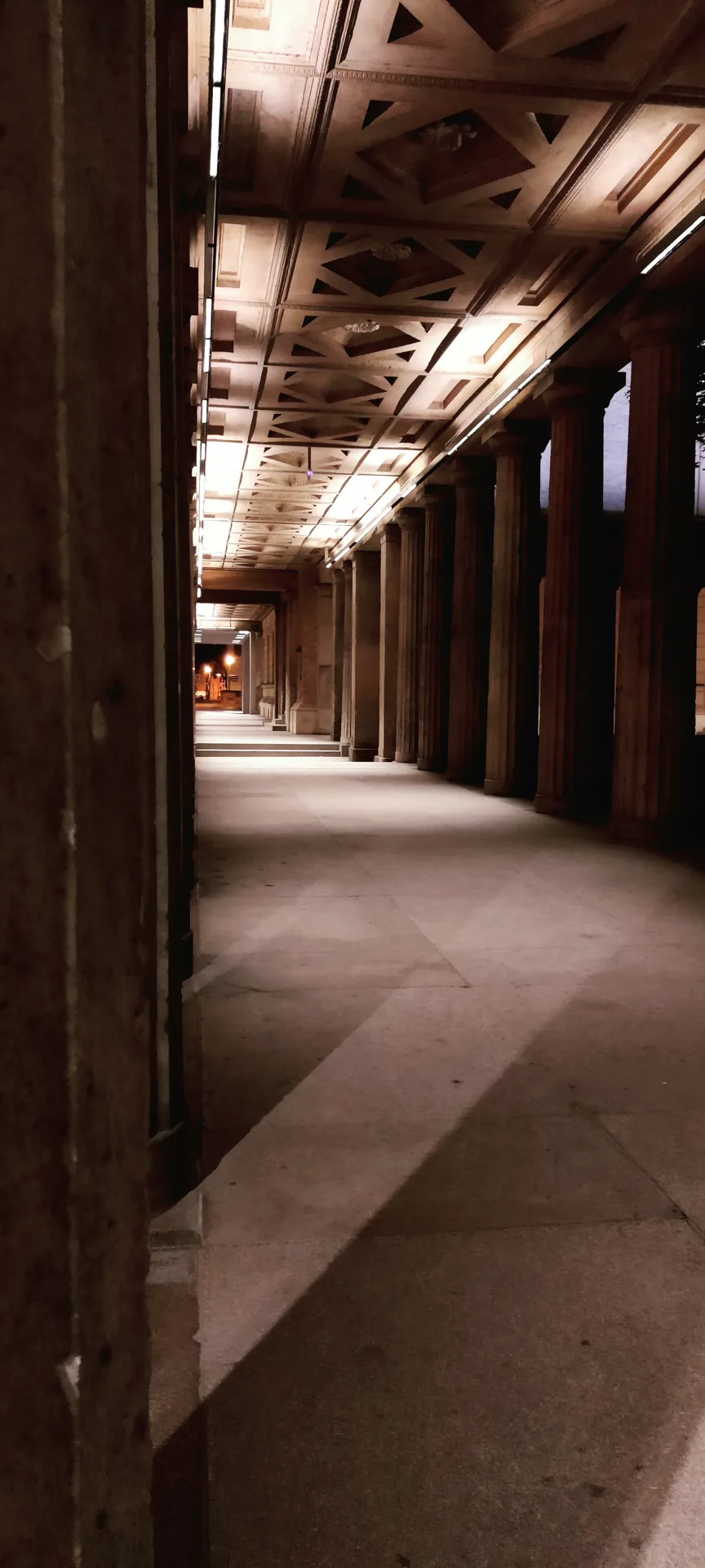 Photo d'une longue colonnade éclairée dans la nuit, son plafond à caissons et son sol vide filant jusqu'à un lampadaire au fond