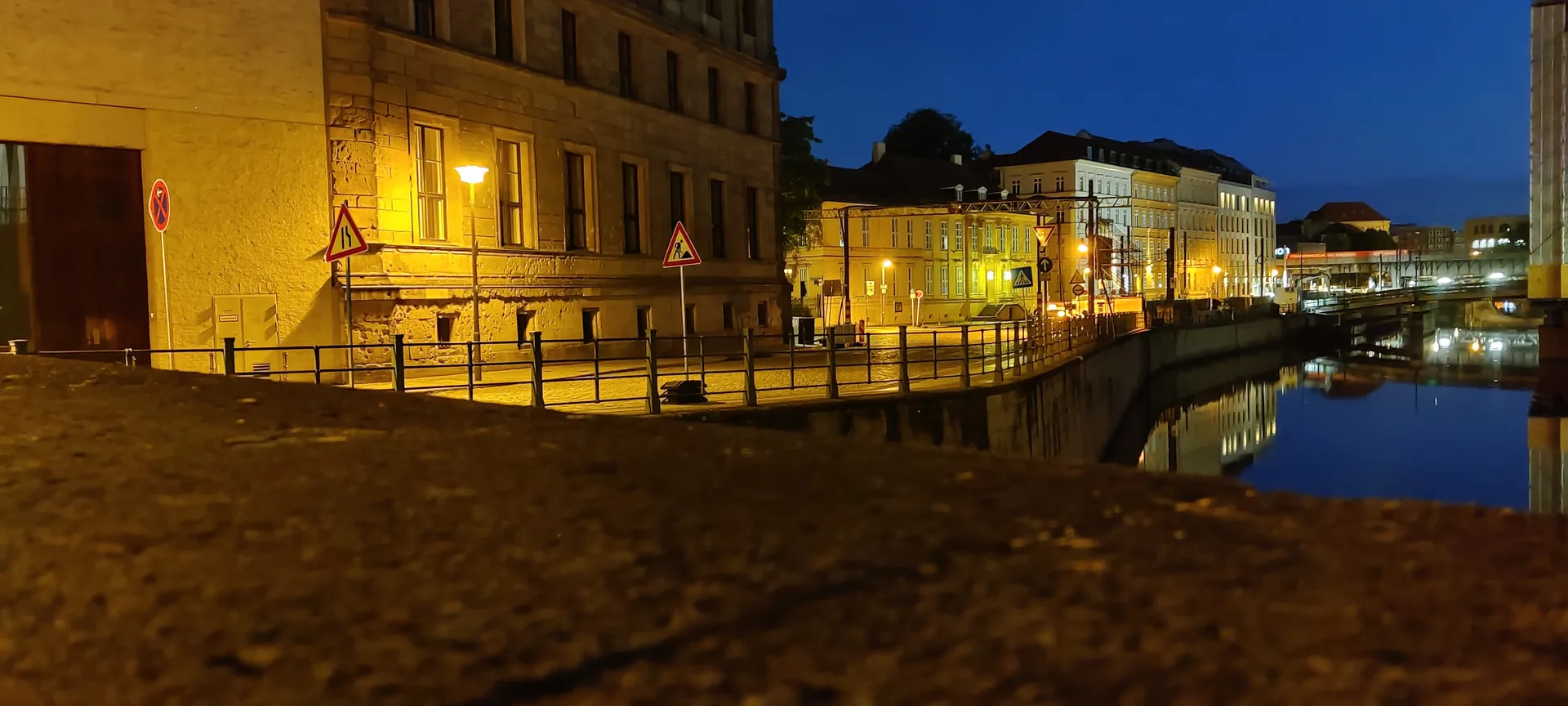 Photo prise au ras d'un parapet de pierre, le quai de la Spree éclairé et ses façades reflétées dans l'eau, personne sur la chaussée