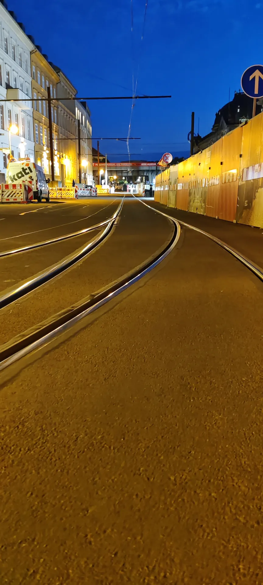 Photo au ras du sol de rails de tramway s'incurvant dans une rue vide, une palissade de chantier à droite et le ciel déjà bleu
